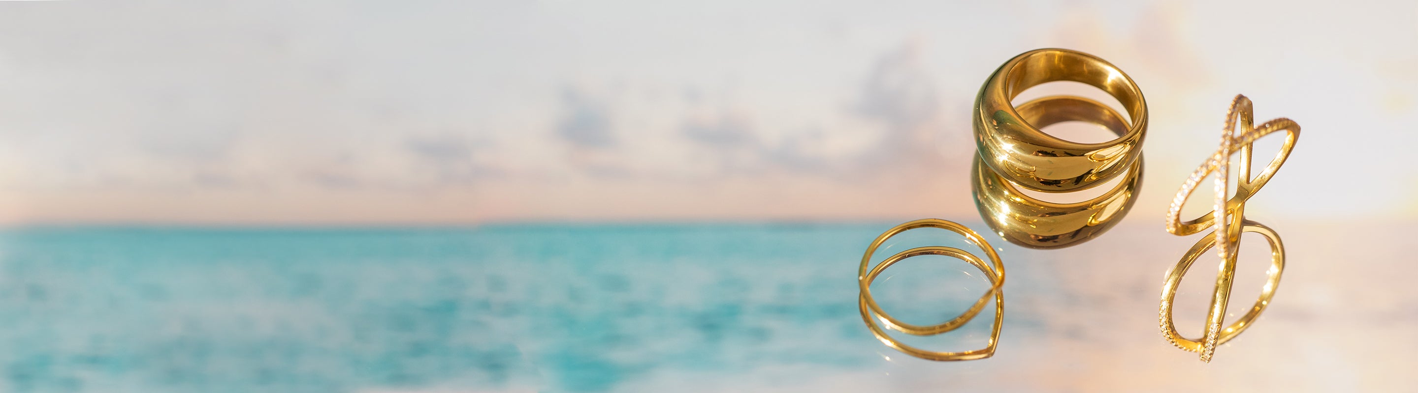 Goldene Ringe von PURELEI, elegant und glänzend, vor einem sanften Ozeanhorizont.