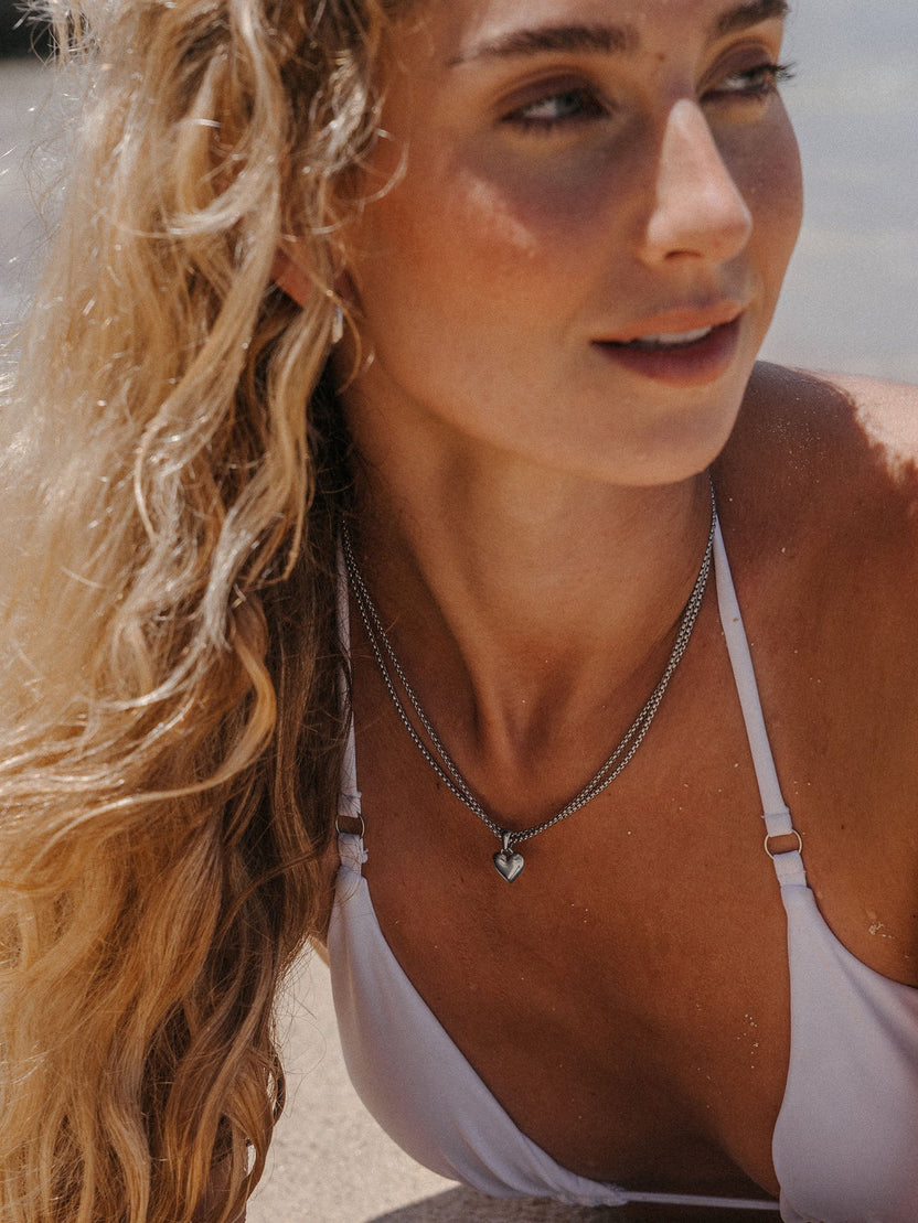 PURELEI Aloha Love Charm Herzanhänger aus silberfarbenem Edelstahl, am Strand getragen, elegant und zeitlos.