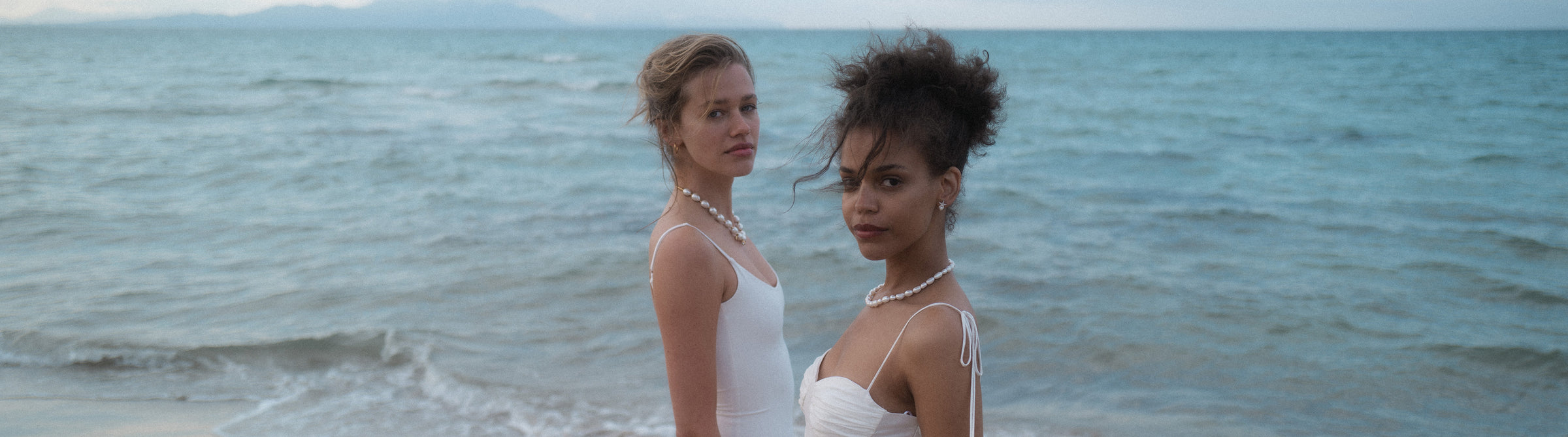Zwei Frauen in weißen Kleidern mit Perlenschmuck am Strand, PURELEI inspiriert.