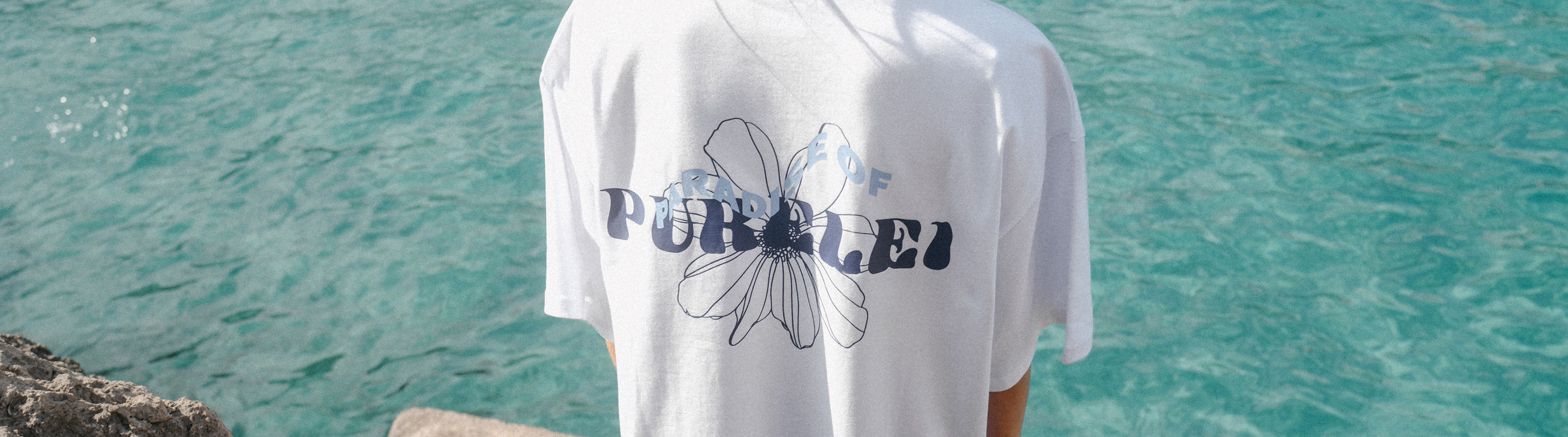 Weißes T-Shirt mit floralem PURELEI-Design, getragen vor türkisfarbenem Wasser.