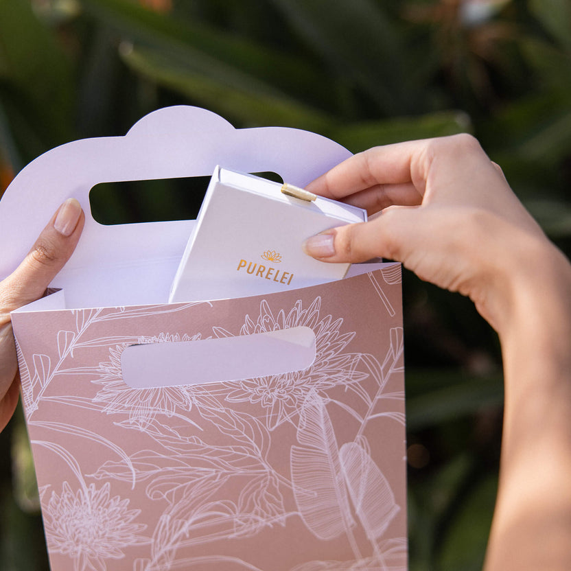 Hände halten eine rosa PURELEI-Tüte mit floralem Design und einer weißen Box darin.