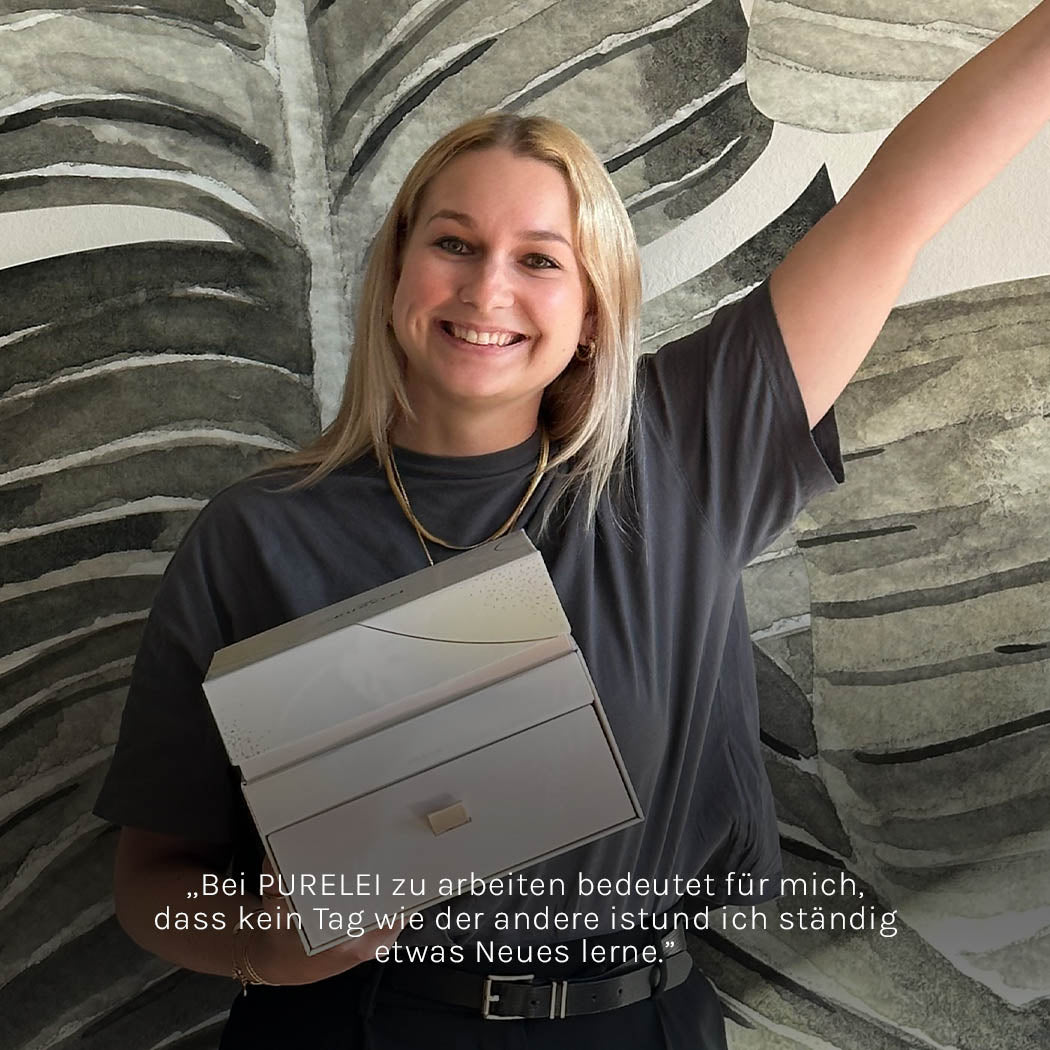 Frau mit lächelndem Gesicht, hält eine PURELEI-Schmuckbox vor tropischem Hintergrund.