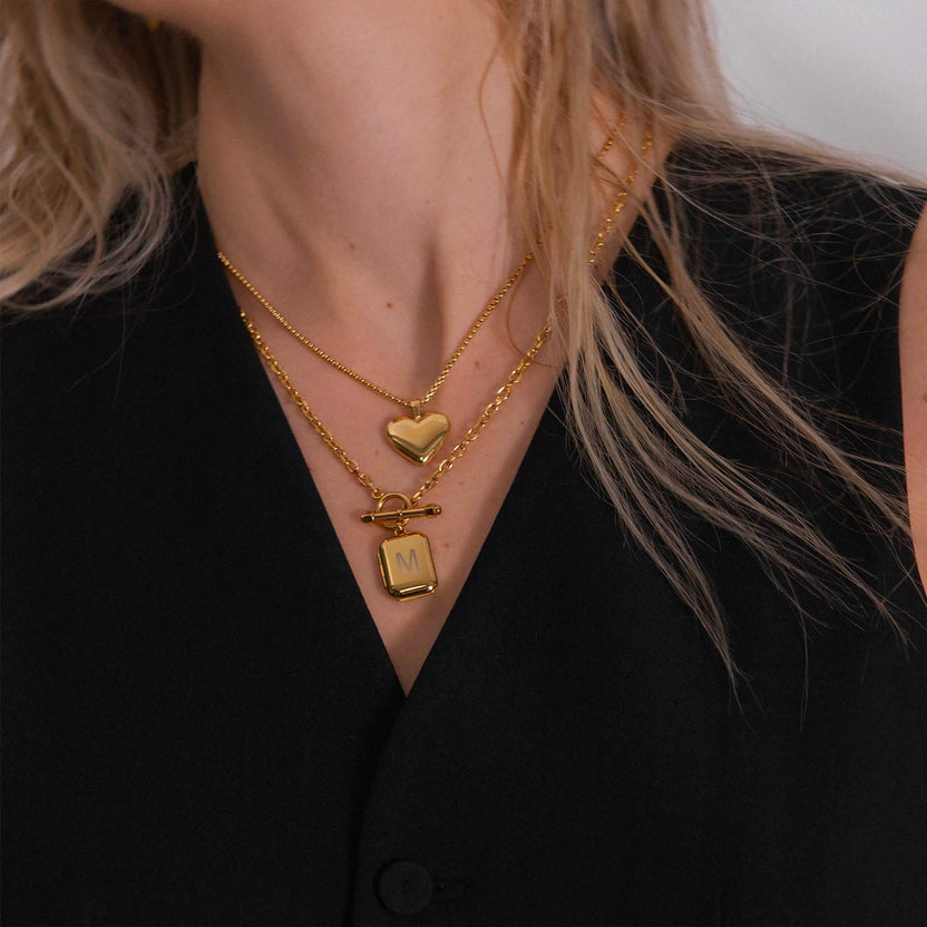 Zwei goldene Ketten mit Herz- und Anhänger-Design, getragen mit schwarzem Oberteil, PURELEI.