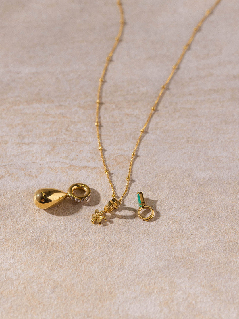 Feingliedrige Li’ili’i Kette in 18K Gold mit zarten Anhängern auf hellem Sand, PURELEI Style, elegant und feminin.