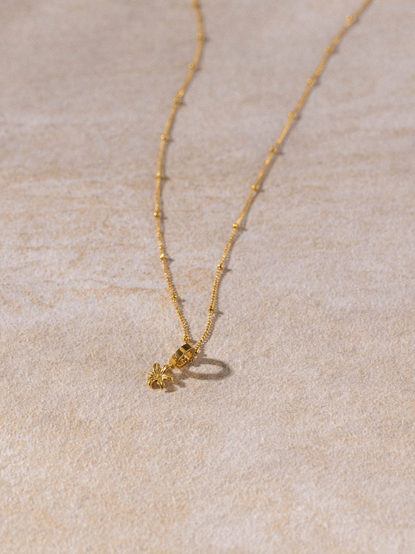 Feingliedrige Li’ili’i Kette aus 18K vergoldetem Edelstahl mit Blumenanhänger auf sandfarbenem Hintergrund PURELEI
