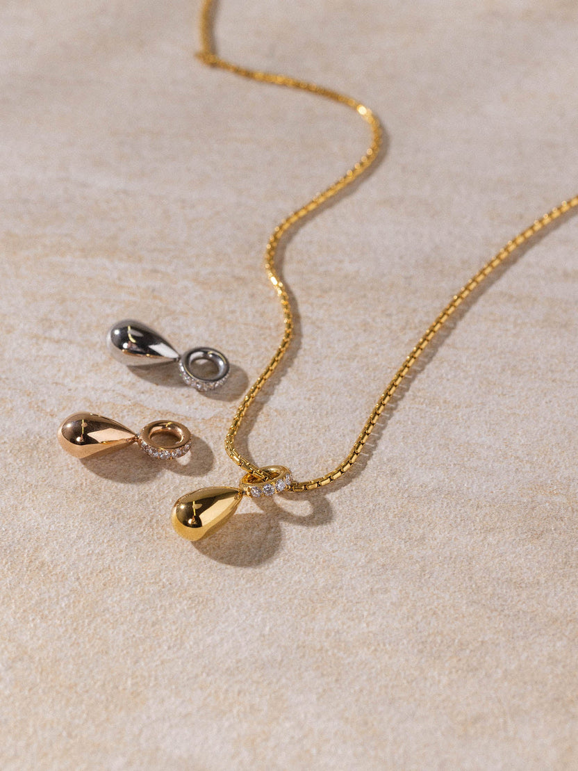 PURELEI Resonance Kette in Gold, Silber und Roségold mit tropfenförmigem Anhänger, clean & elegant auf sandfarbenem Hintergrund
