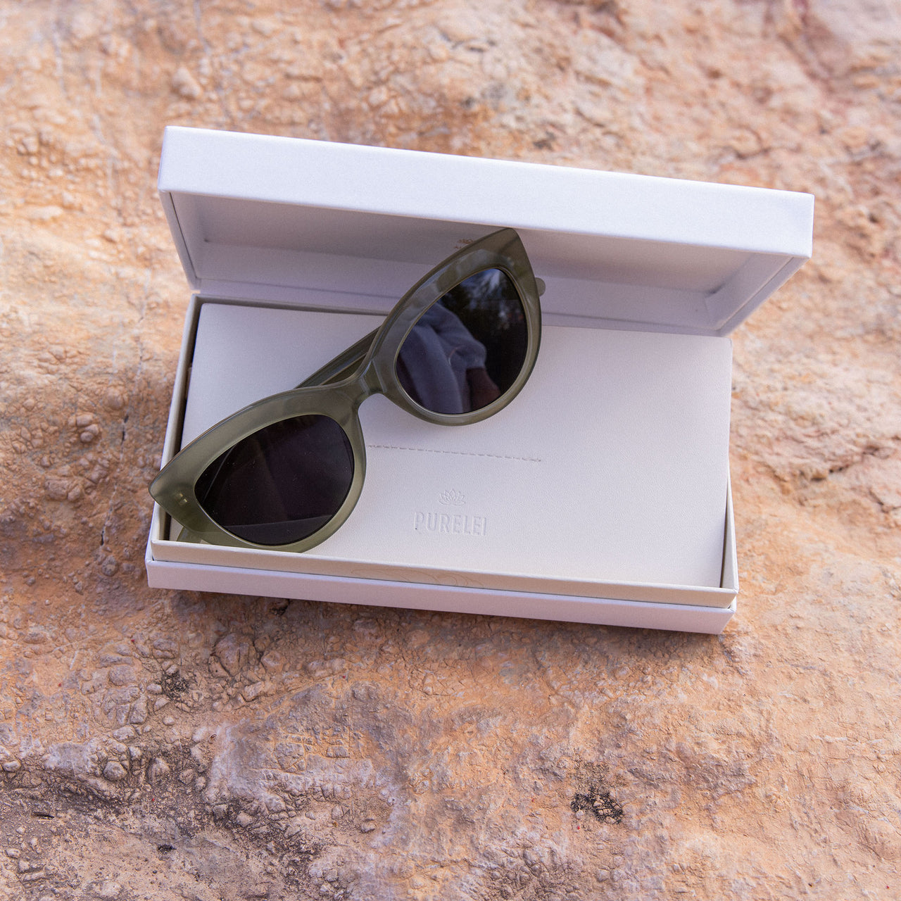 Stylische, grüne Cat-Eye-Sonnenbrille in PURELEI Box auf natürlichem Untergrund.