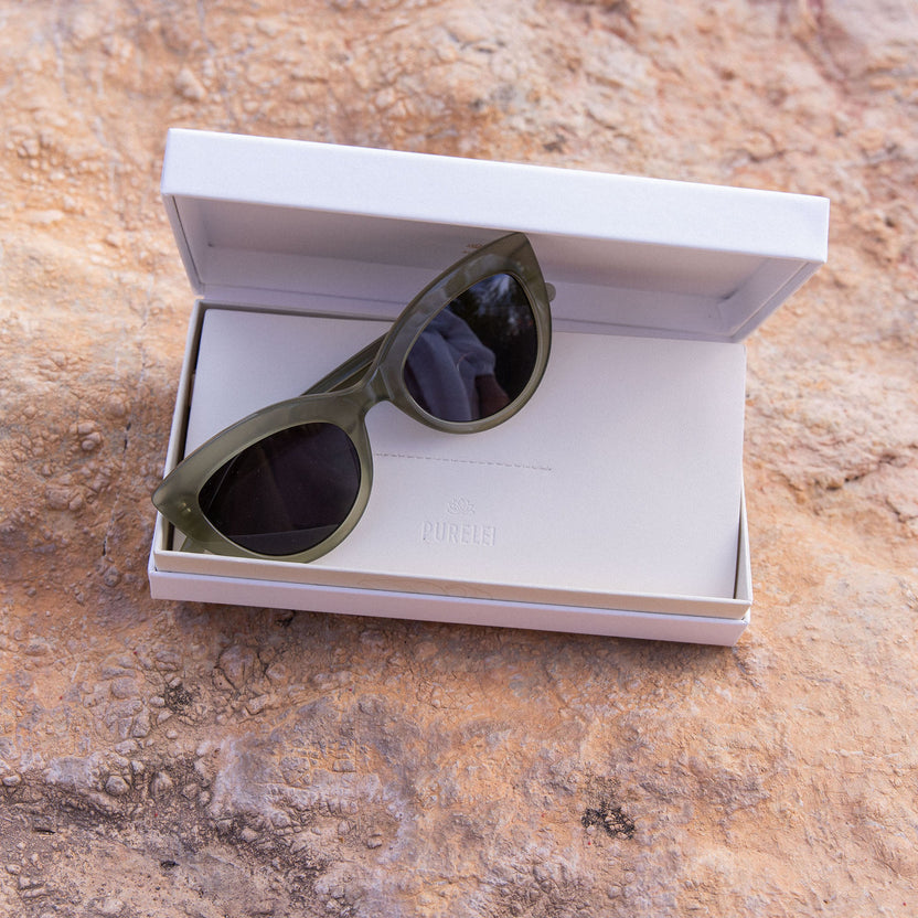 Stylische, grüne Cat-Eye-Sonnenbrille in PURELEI Box auf natürlichem Untergrund.