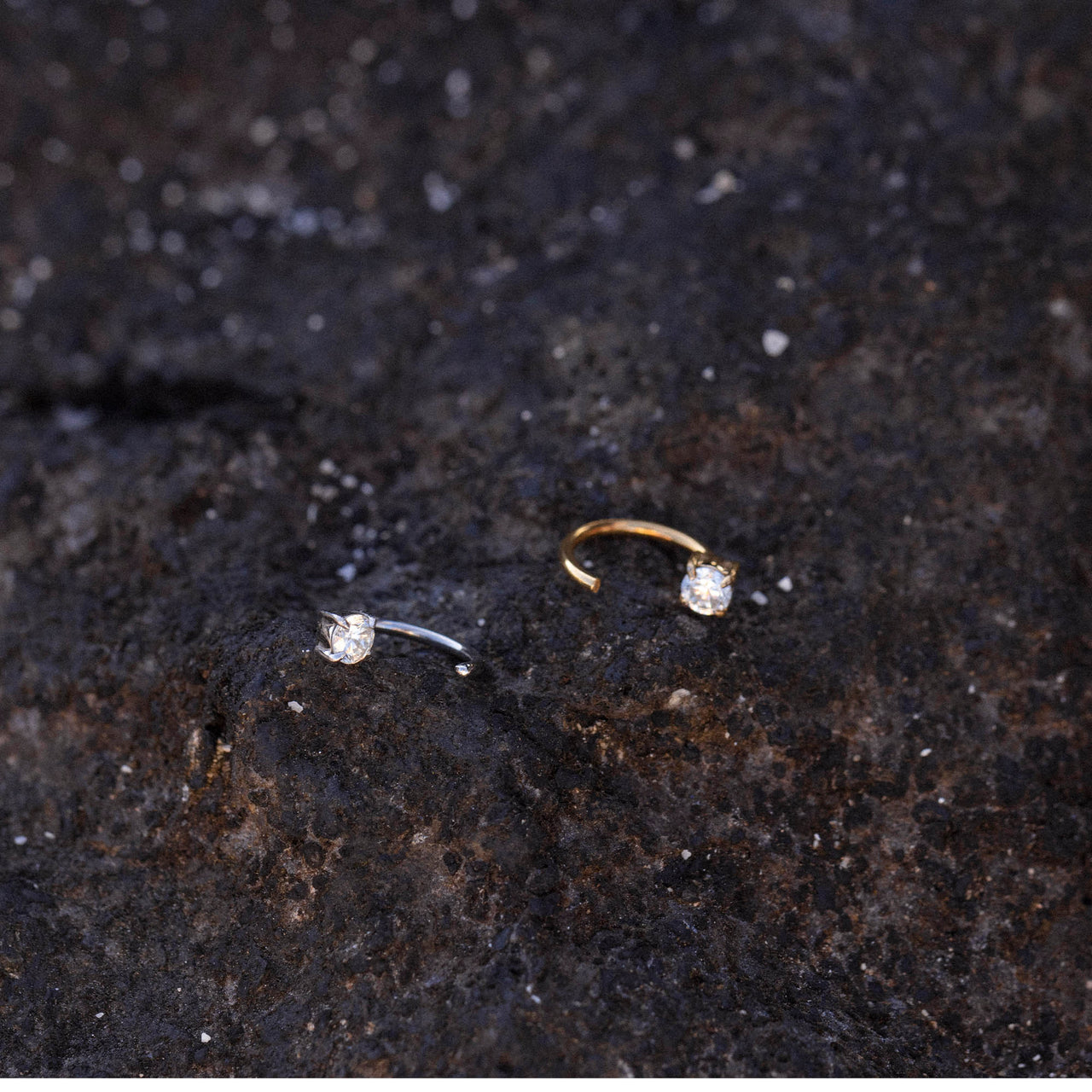Highlight Piercing von PURELEI, gold- und silberfarbene Edelstahlringe mit funkelndem Zirkonia auf dunklem Lavagestein.