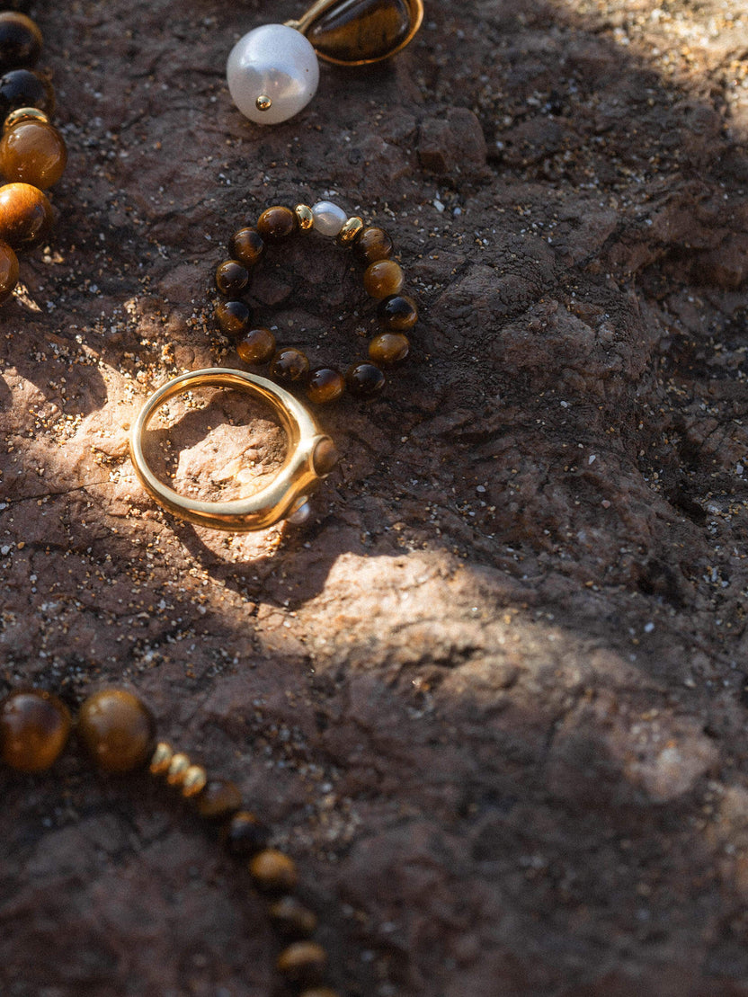 Elastic Anxiety Tiger‘s Eye Pearl Ring mit goldbraunen Tigeraugeperlen und zarter Süßwasserperle auf dunklem Stein.