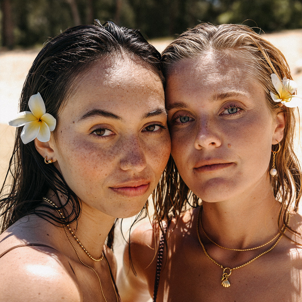 Zwei Frauen mit PURELEI Schmuck, Muschel-Anhänger und Blumen im Haar, am Strand.