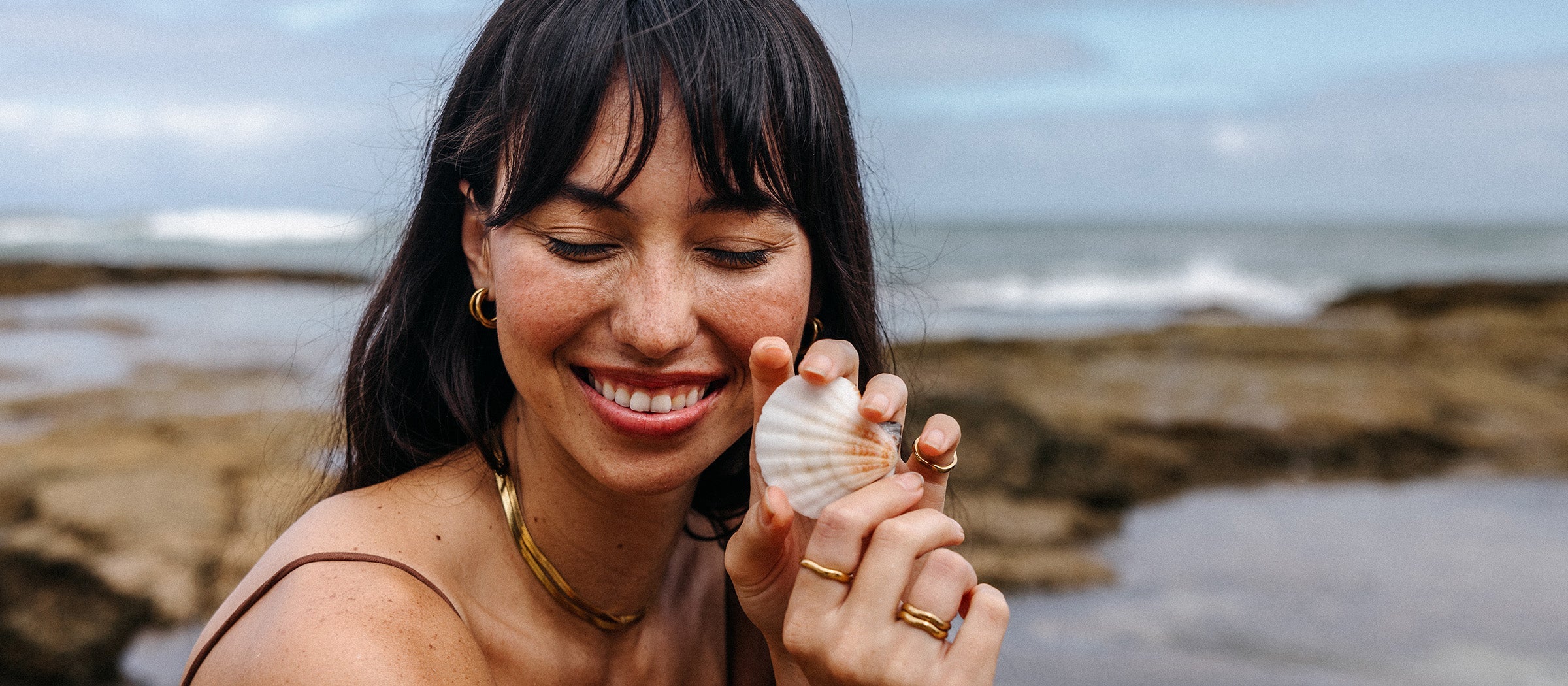 Frau mit Muschel und PURELEI Schmuck am Strand, natürliche Farben und fröhliche Stimmung.