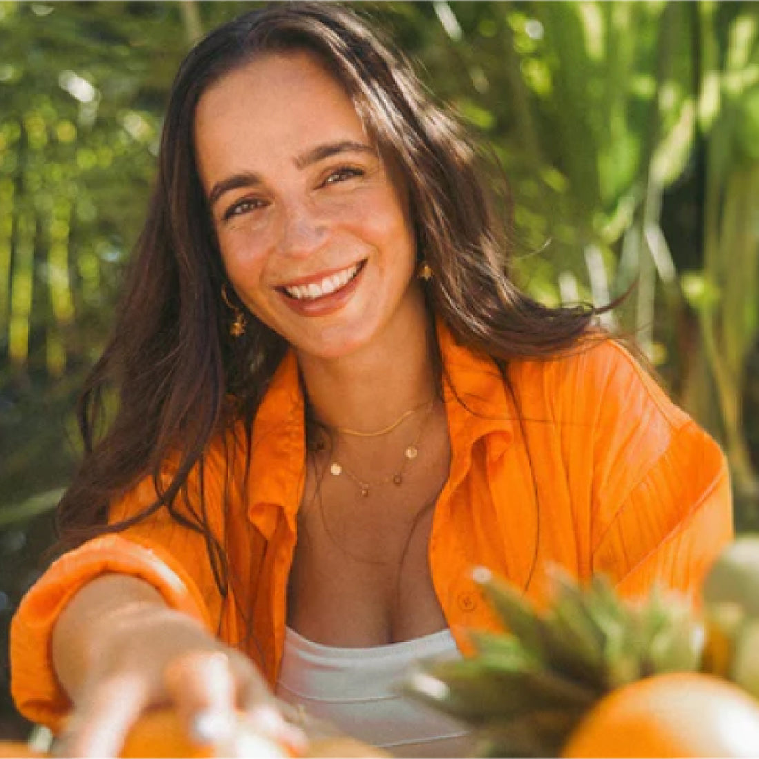 Frau mit orangefarbenem Shirt und PURELEI Schmuck, umgeben von tropischer Natur.