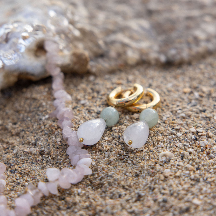 Zarte PURELEI Ohrringe und Kette aus gold und weißen Steinen auf Sand.