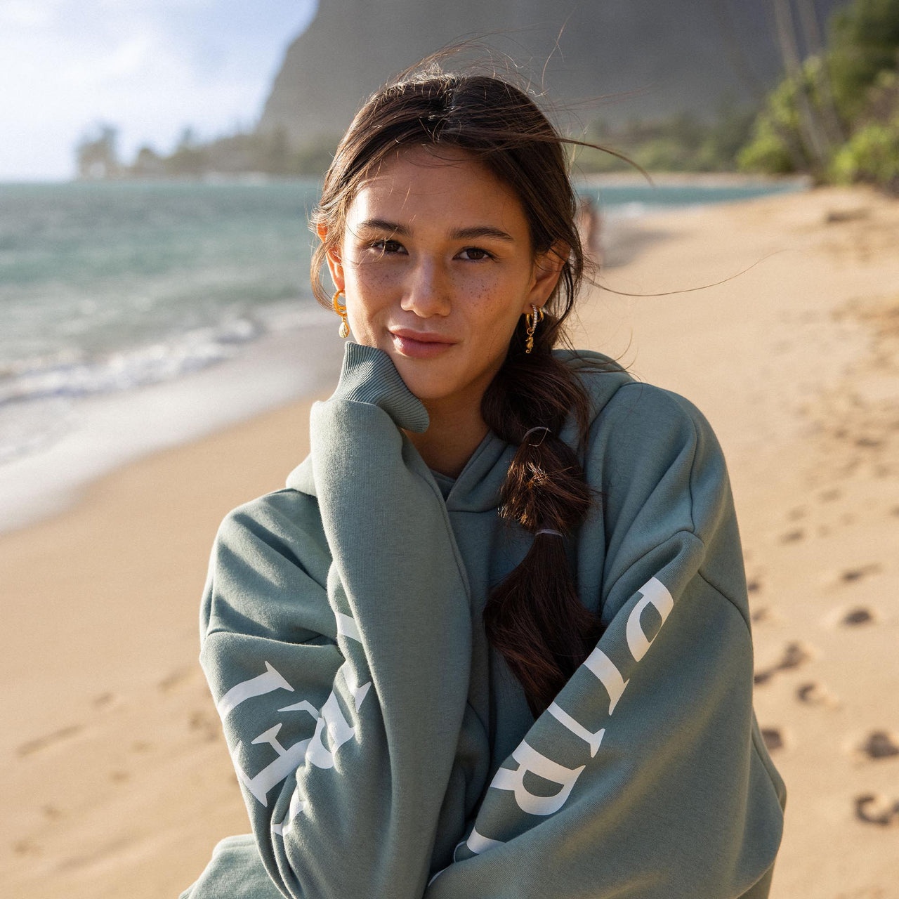 Junge Frau in grauem Hoodie am Strand, PURELEI inspiriert von Aloha-Vibes.